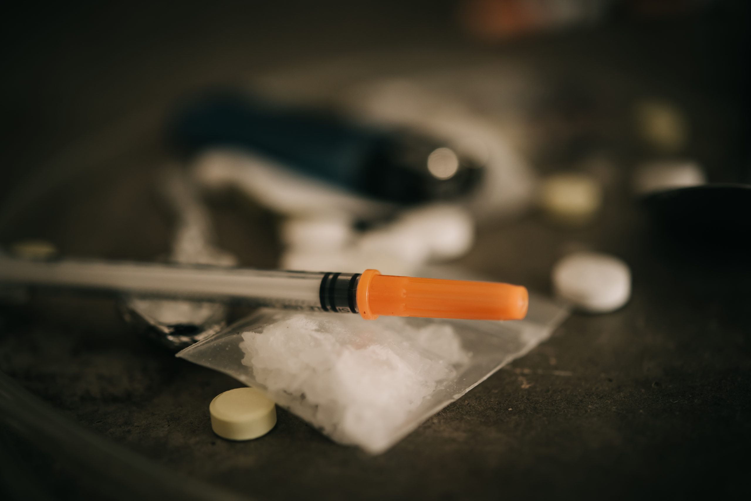 Syringe with orange cap resting on a plastic bag of white powder, surrounded by scattered pills and a lighter.