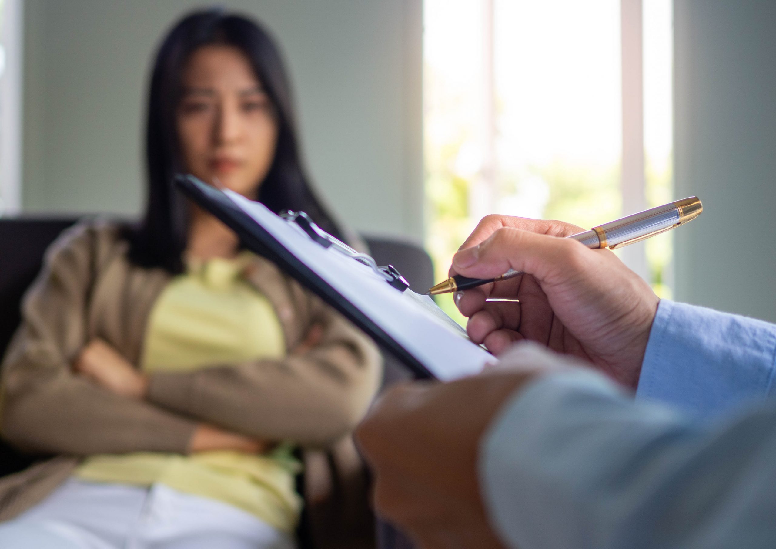 A therapist holds a clipboard and pen while a woman sits on a couch, looking contemplative with arms crossed.