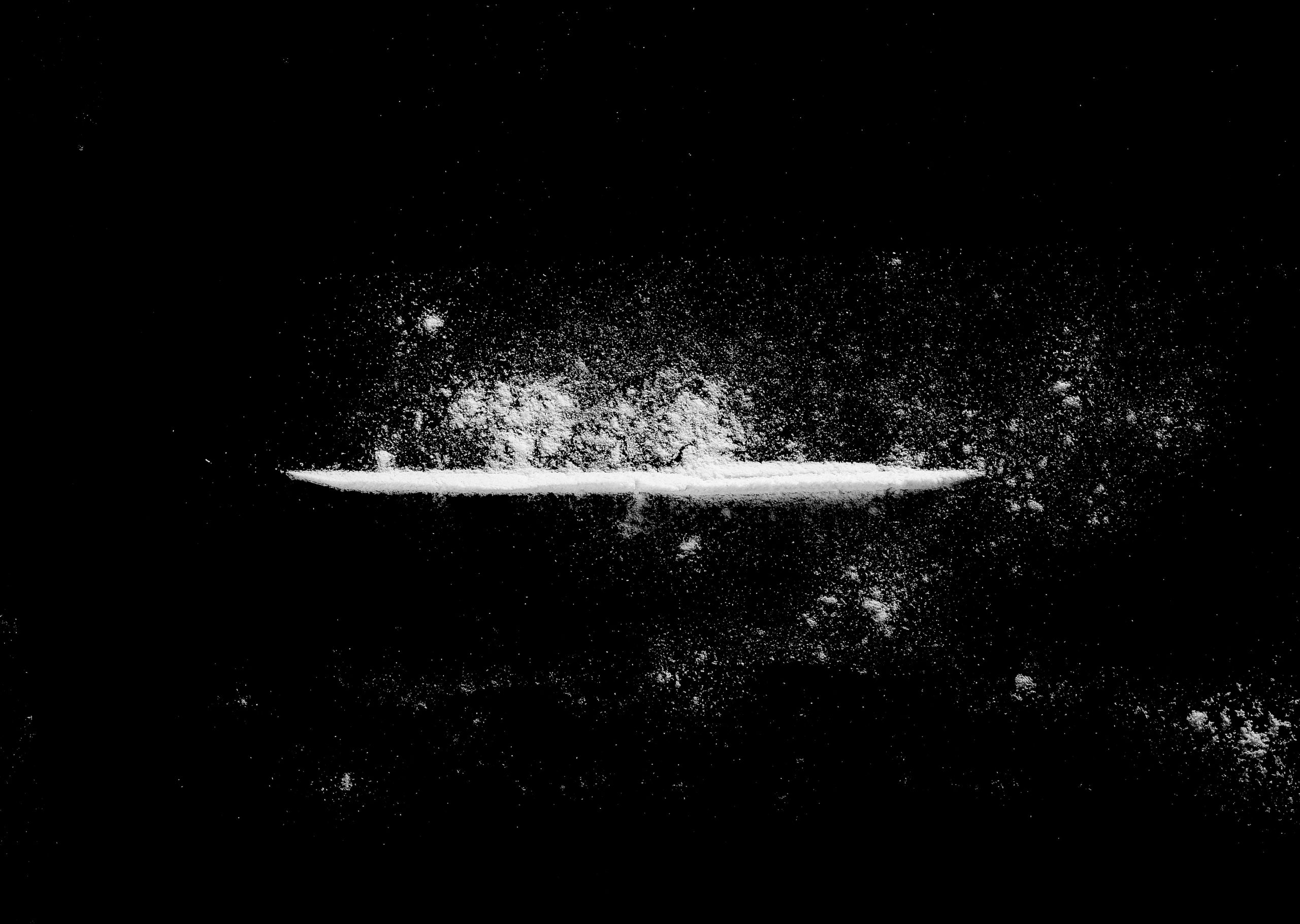 A thin line of white powder against a black background, with scattered particles creating a textured effect.