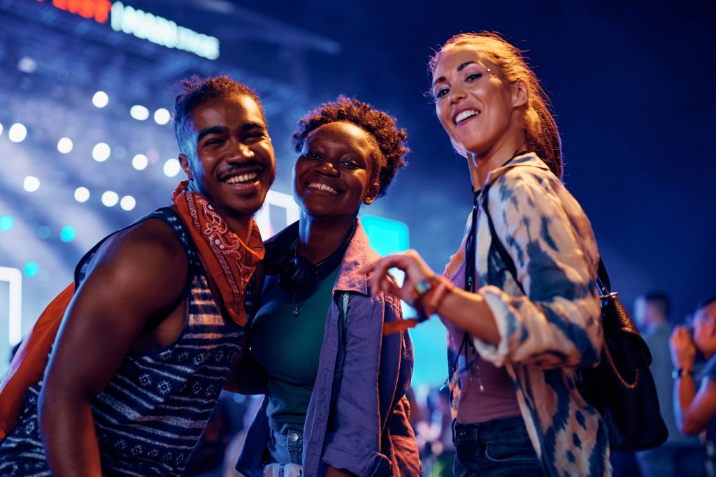 Three friends smiling and posing together at a vibrant concert, colorful lights in the background.