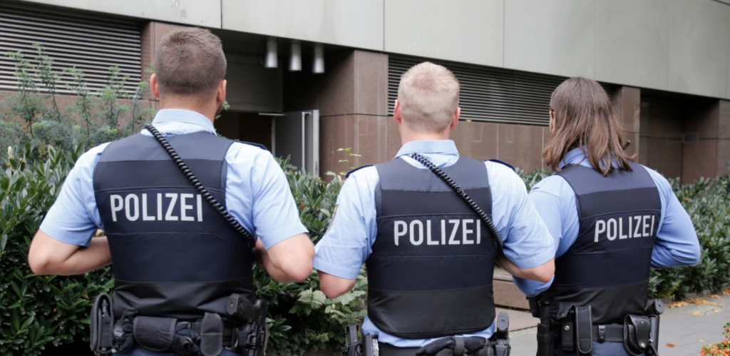 Three police officers in blue uniforms, backs turned, walking towards a building surrounded by greenery.