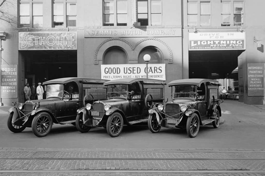 Three vintage cars parked in front of the Semmes Motor Company, with signs advertising "Good Used Cars" and "Lightning Motor.