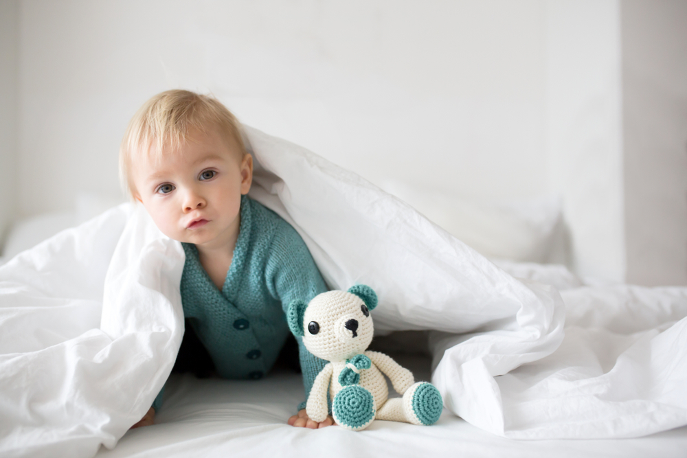 Toddler peeking out from under a white blanket, beside a crocheted bear toy, in a cozy bedroom setting.