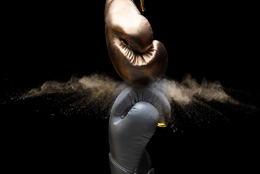Two boxing gloves collide, one gold and one black, with a cloud of dust bursting between them against a dark background.