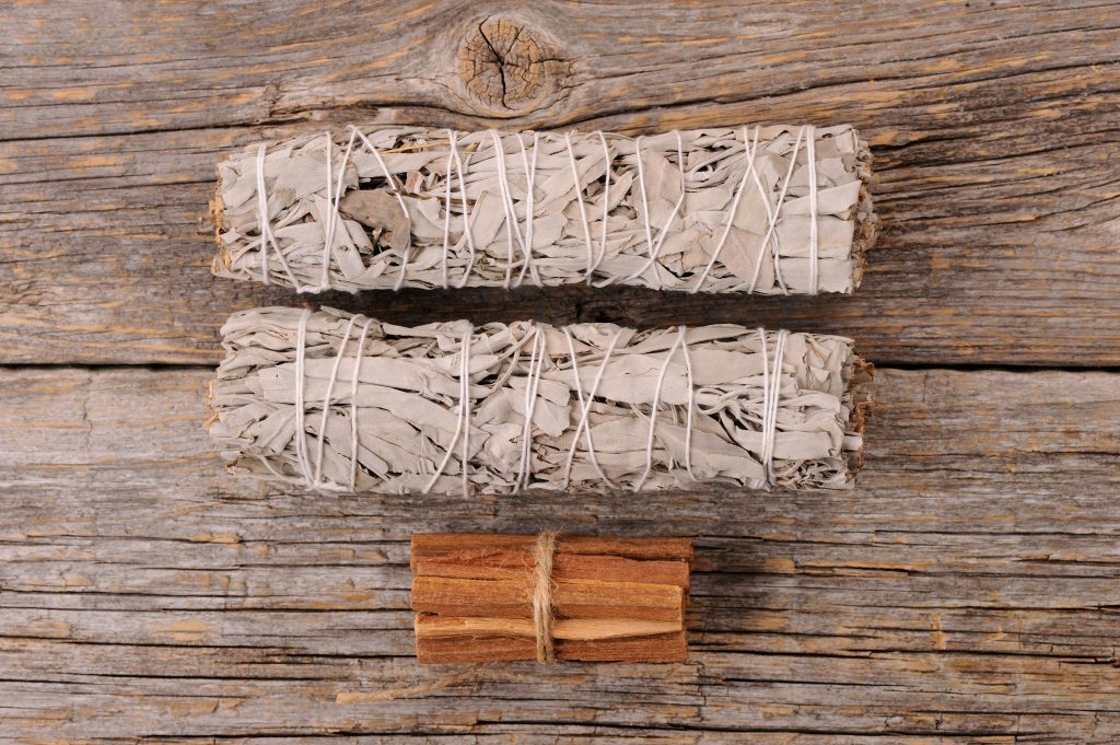 Two bundles of white sage tied with string, alongside a smaller bundle of brown wood, resting on a rustic wooden surface.