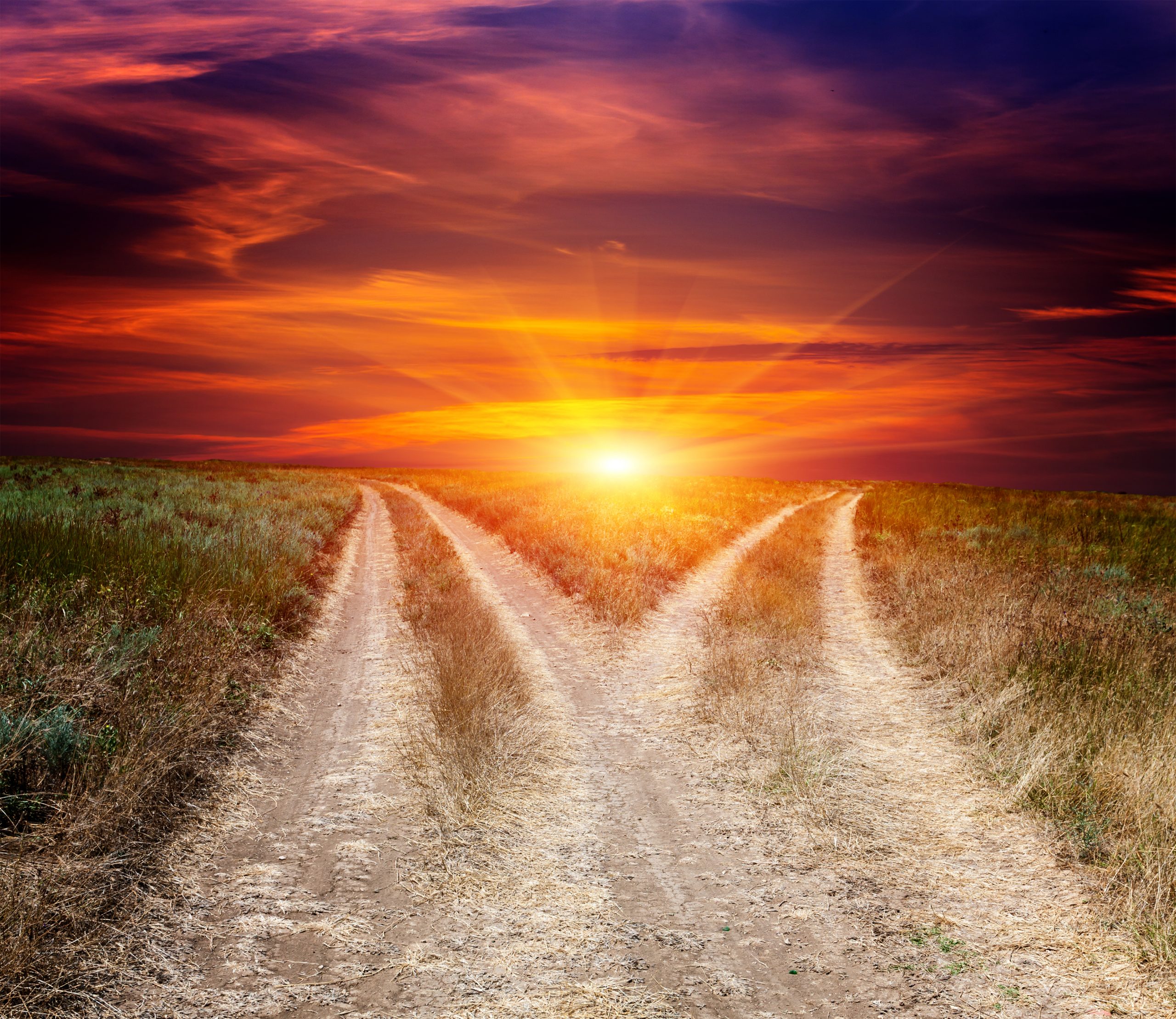 Two diverging dirt paths lead towards a vibrant sunset, with colorful clouds illuminating the sky.