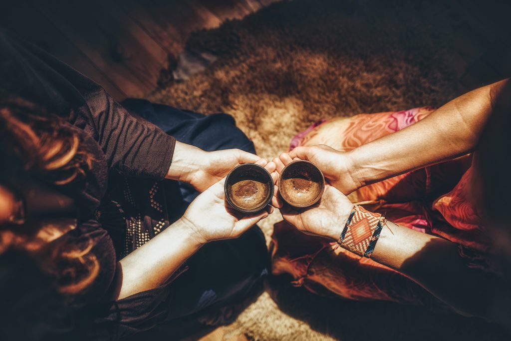 Two hands hold small bowls, sharing a warm drink, surrounded by a cozy, softly lit setting.
