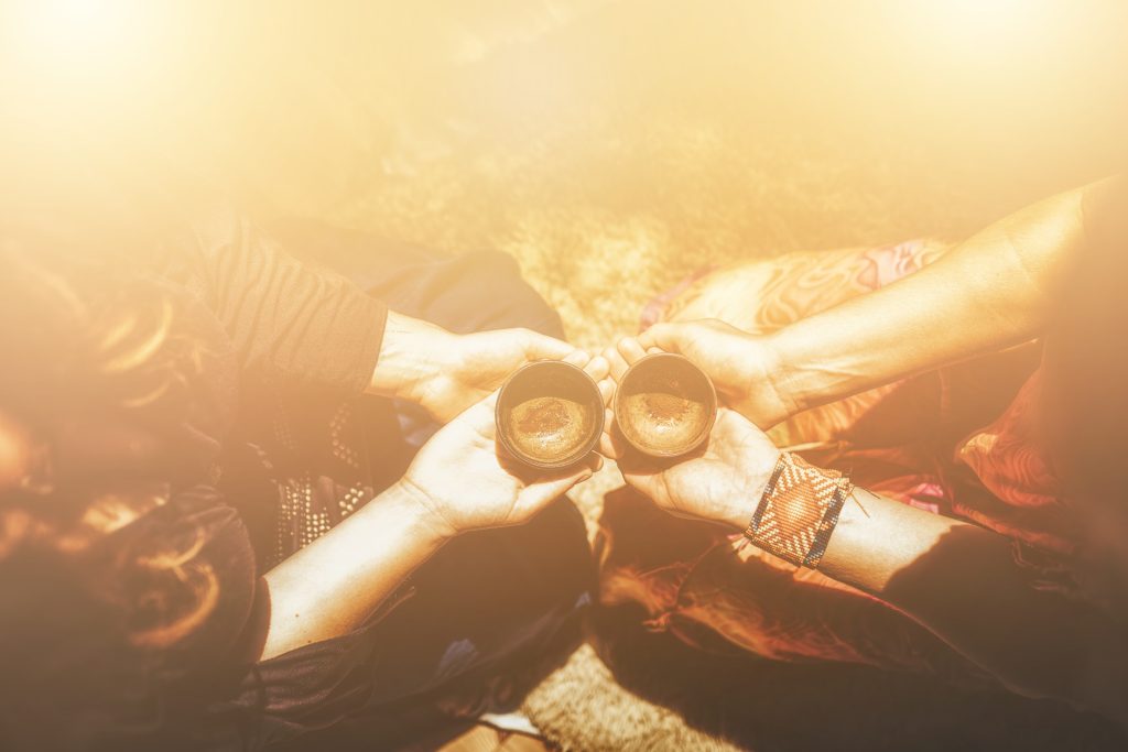 Two hands hold small clay cups, sharing a warm beverage, with sunlight softly illuminating the scene.