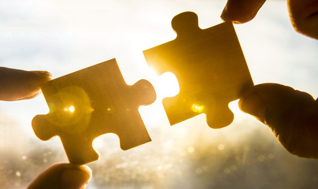 Two hands hold wooden puzzle pieces, connecting them against a bright, glowing background.