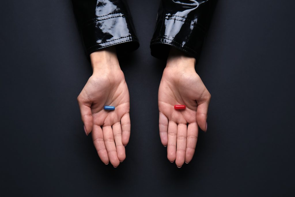 Two hands open, one holding a blue capsule and the other a red capsule, set against a dark background.