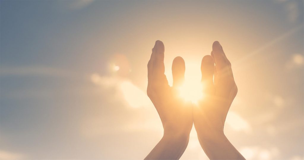 Two hands raised toward the sky, framing a bright sun against a soft, cloudy backdrop.