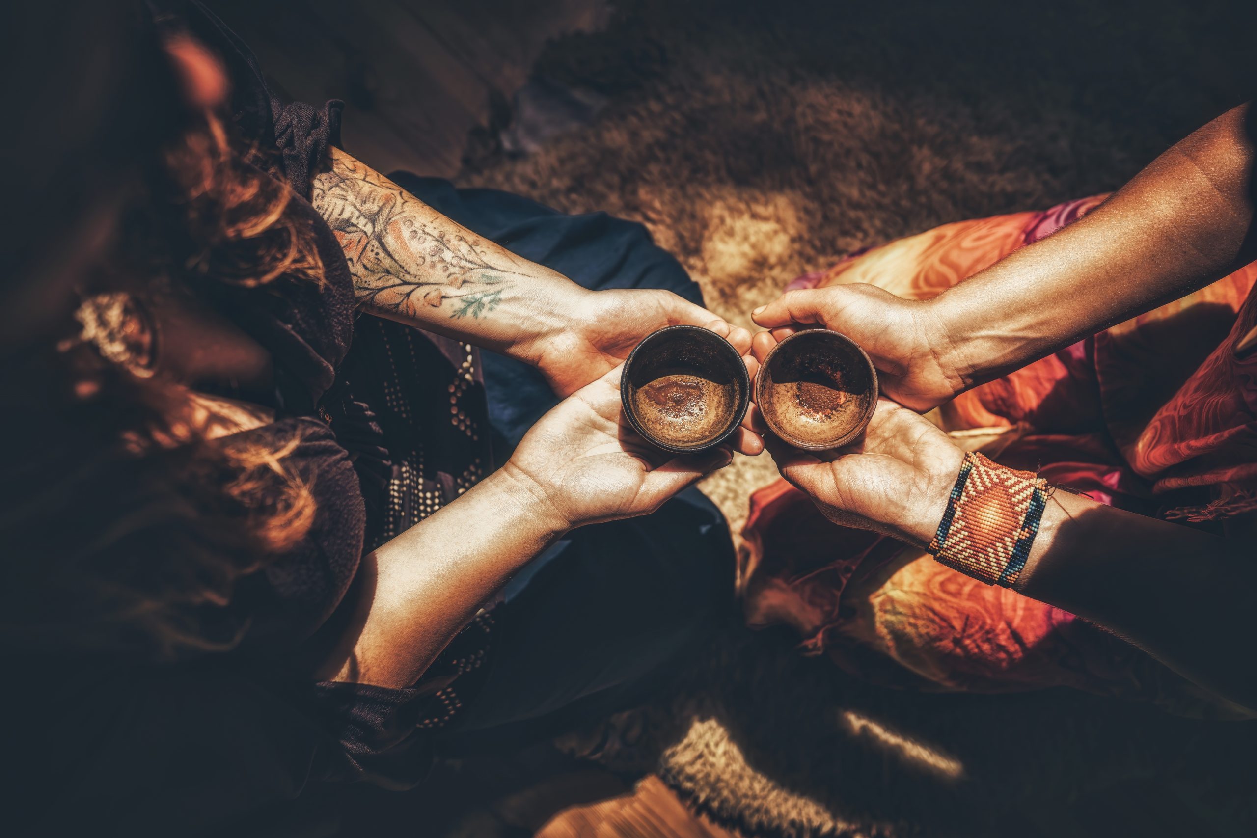 Two individuals hold small, dark bowls in their hands, seated on a textured surface, with warm, earthy tones surrounding t...