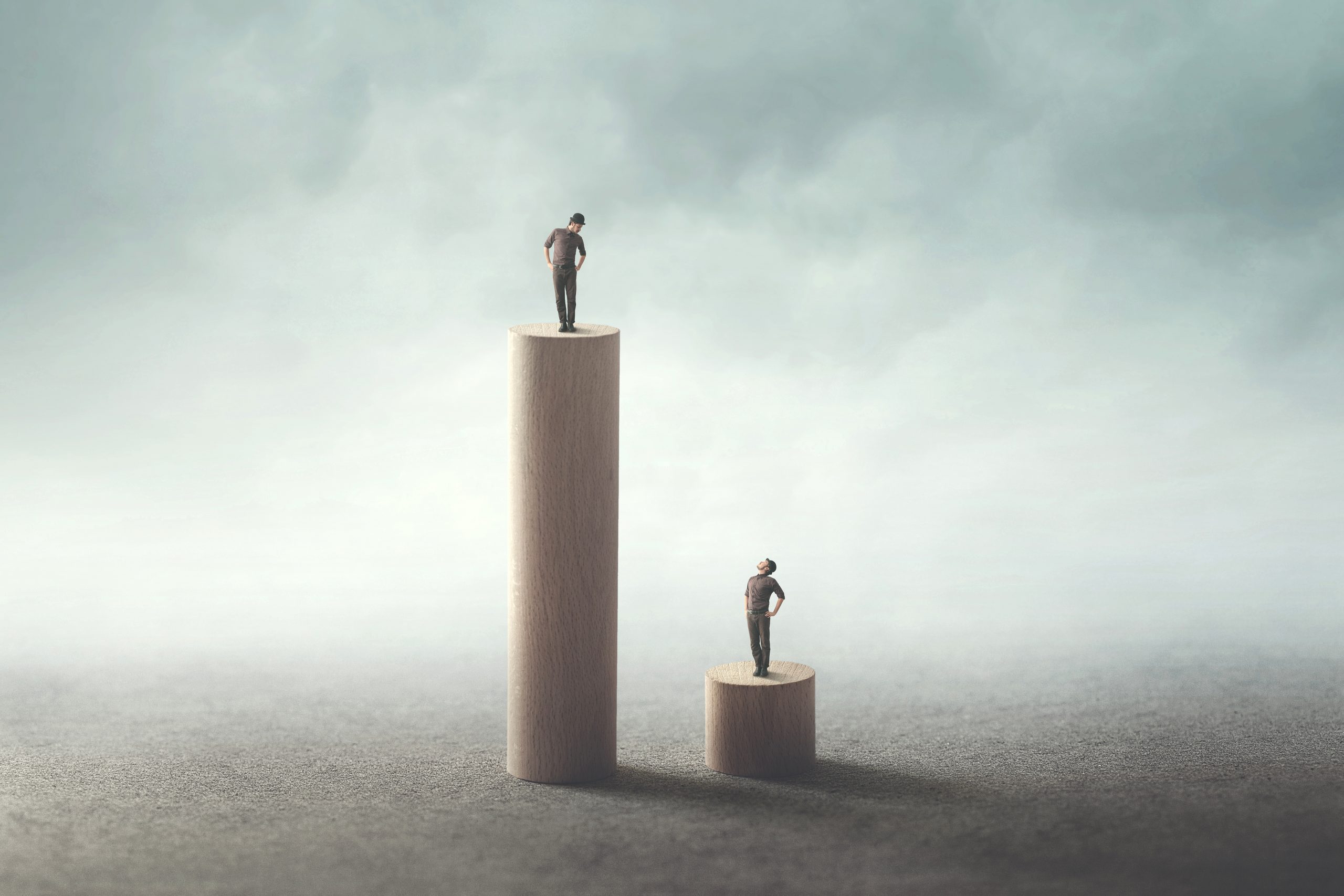Two miniature figures stand atop wooden cylinders of varying heights, symbolizing disparity or achievement against a misty...