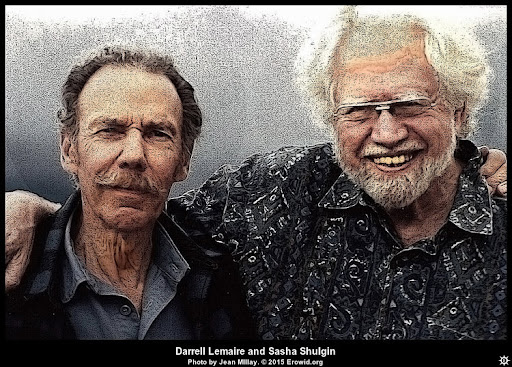 Two older men stand closely together, smiling, with one wearing glasses and a patterned shirt against a blurred background.
