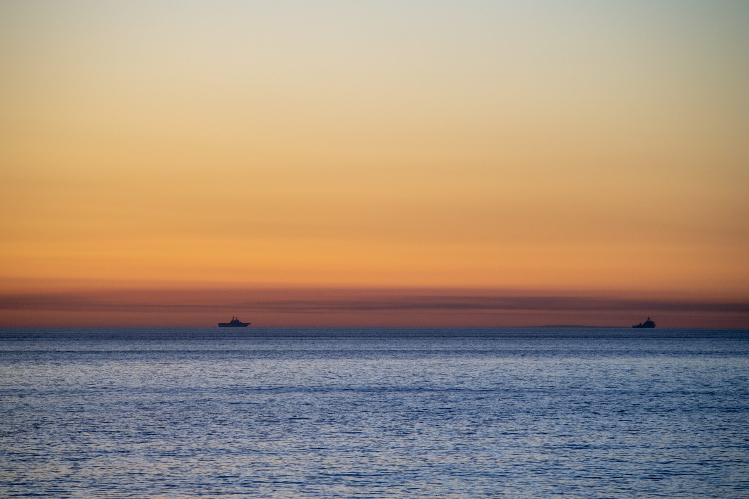 Two ships silhouette against a serene sunset, with soft orange and blue hues reflecting on calm waters.