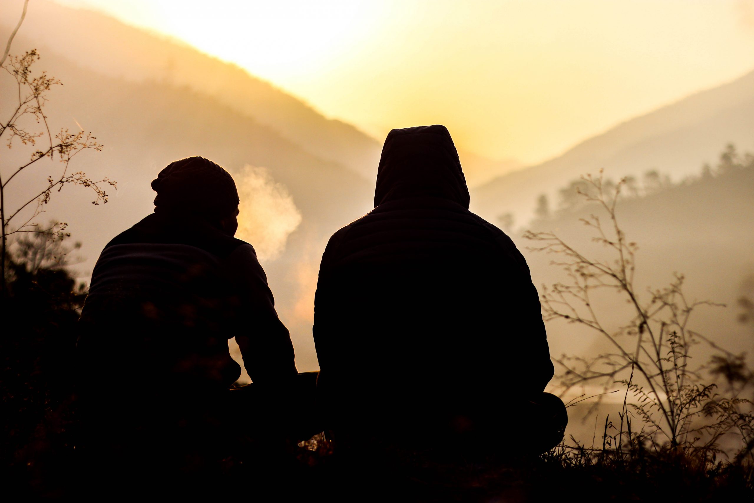 Two silhouetted figures sit together, surrounded by misty mountains at sunset, with one exhaling smoke.