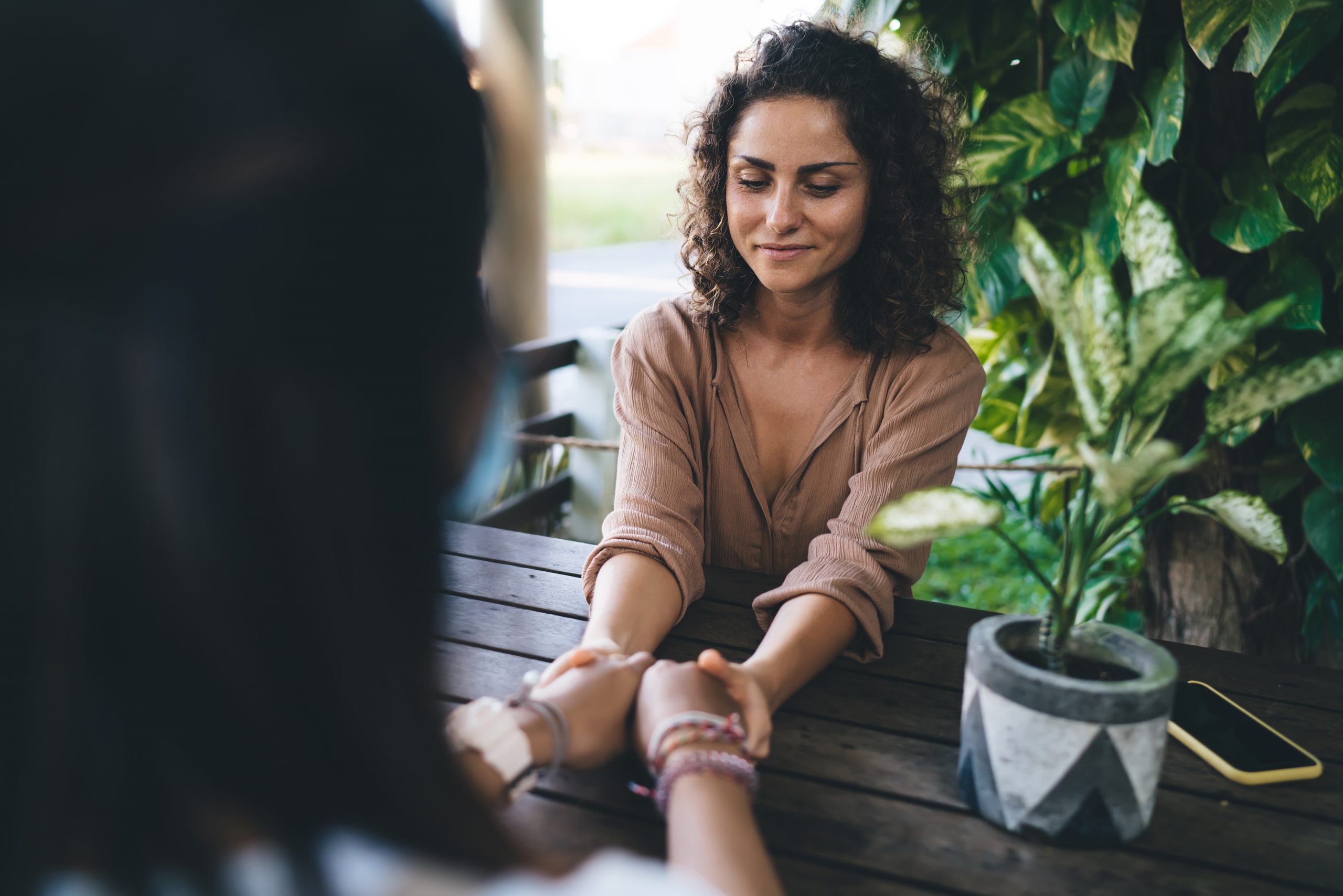 Two women hold hands across a wooden table, surrounded by greenery and a potted plant, sharing a moment of connection.