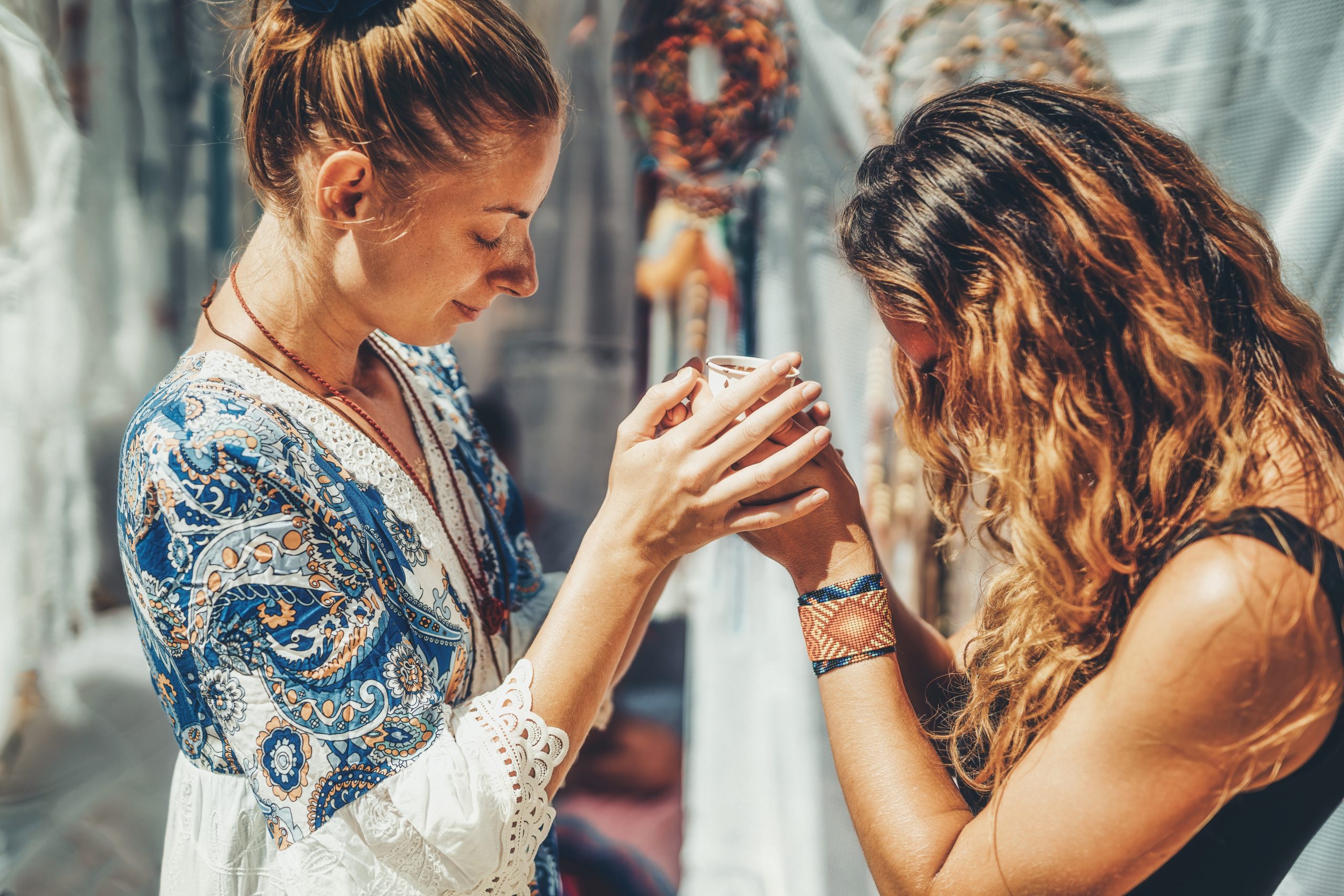 Two women share a moment, holding a small cup, surrounded by dreamcatchers and soft, flowing fabrics.