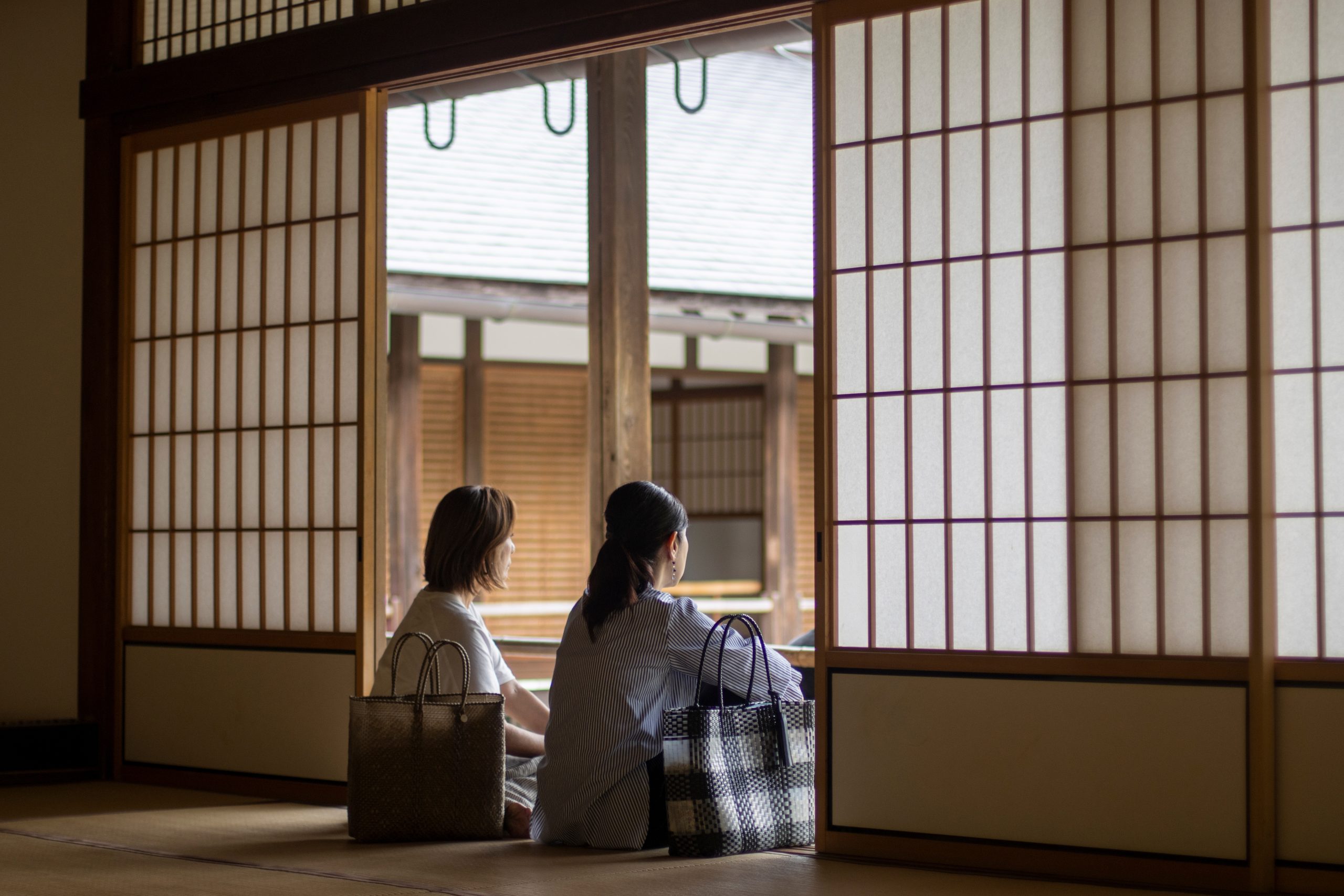 Two women sit on a tatami mat, gazing out through shoji screens, with bags beside them in a serene, traditional setting.