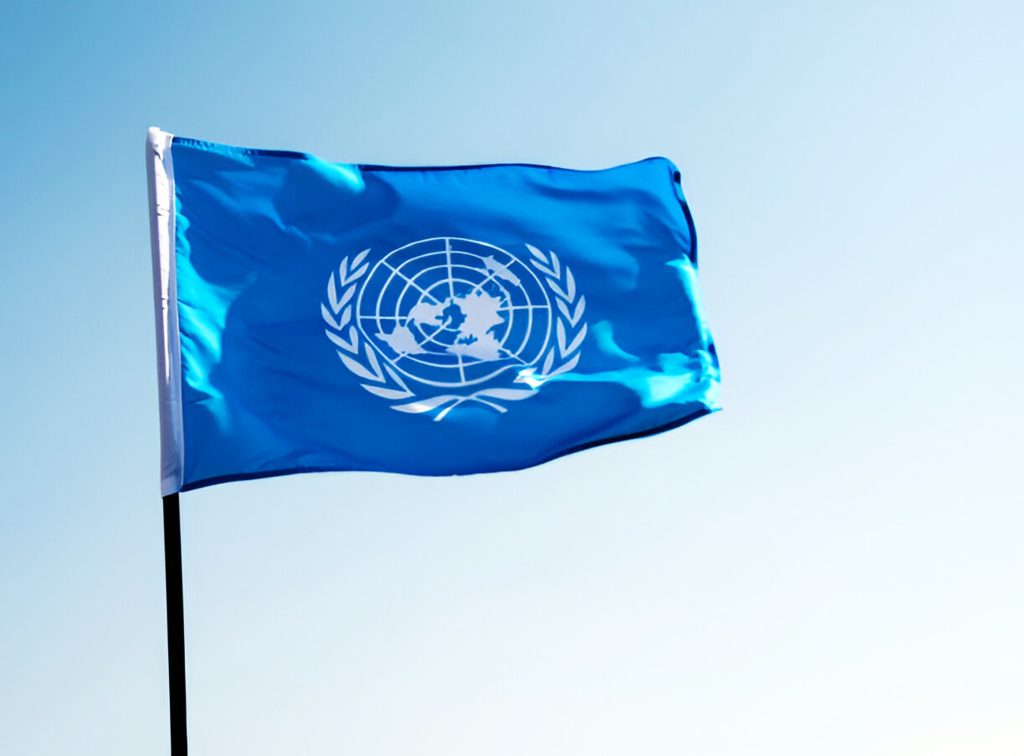 United Nations flag waving against a clear blue sky, featuring the emblem of the globe surrounded by olive branches.