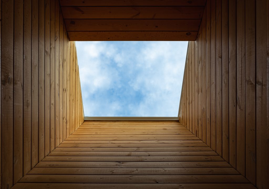 View of a square opening framed by wooden walls, revealing a blue sky with scattered clouds above.