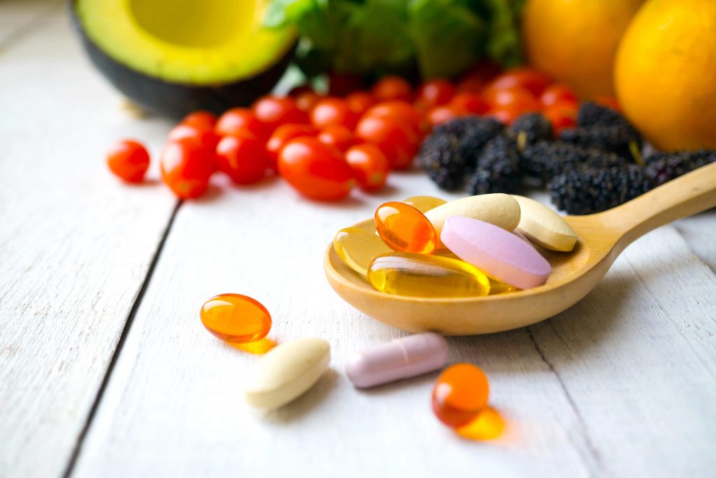 Vitamins and supplements on a wooden spoon, surrounded by fresh fruits and vegetables, including avocado and tomatoes.