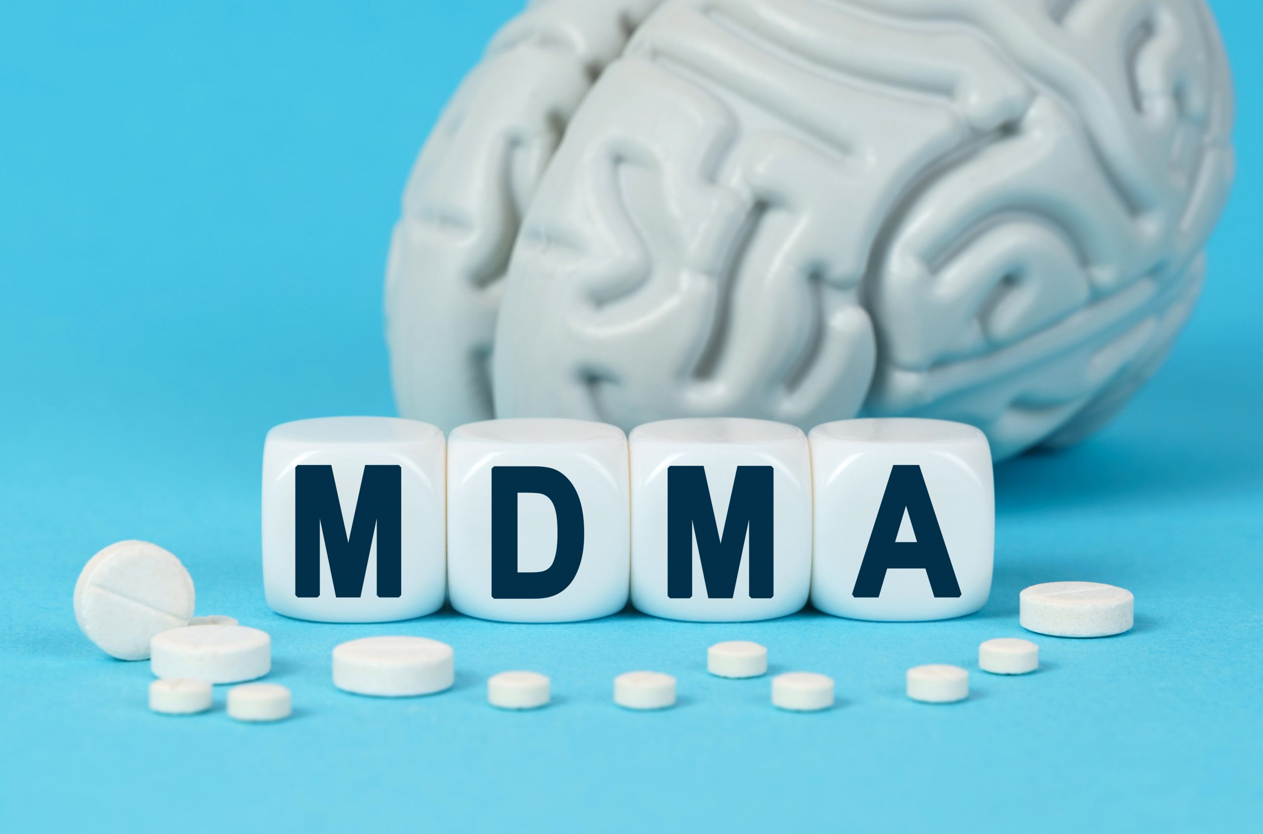 White blocks spell "MDMA" in front of a gray brain model and scattered white pills on a blue background.