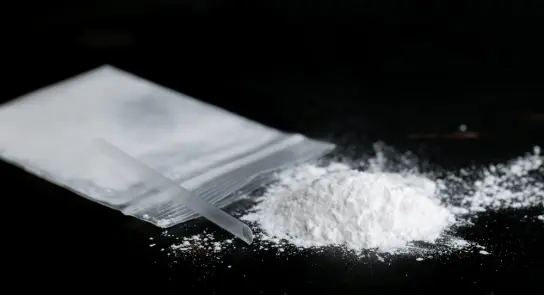 White powder spilled from a clear plastic bag, with a small straw beside it on a dark surface.