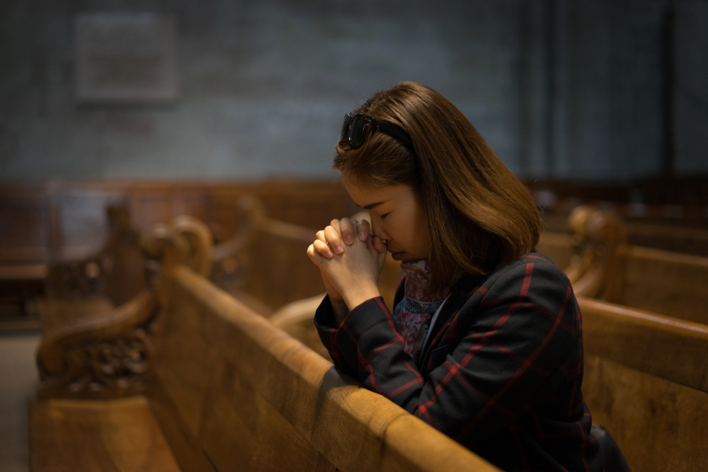 A woman with brown hair prays quietly in a wooden church pew, surrounded by a serene, dimly lit interior.