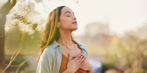 Woman with closed eyes, hands on her chest, enjoying a serene moment outdoors in soft sunlight.