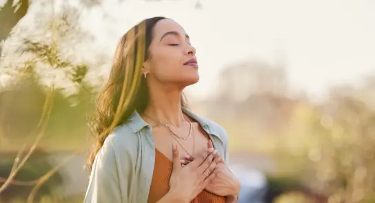 Woman with closed eyes, hands on her chest, enjoying a serene moment outdoors in soft sunlight.