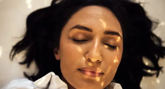 A woman with closed eyes lies on a light surface, soft sunlight casting patterns on her face.