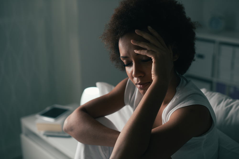A woman with curly hair sits on a bed, looking distressed, with her hand on her forehead in a dimly lit room.