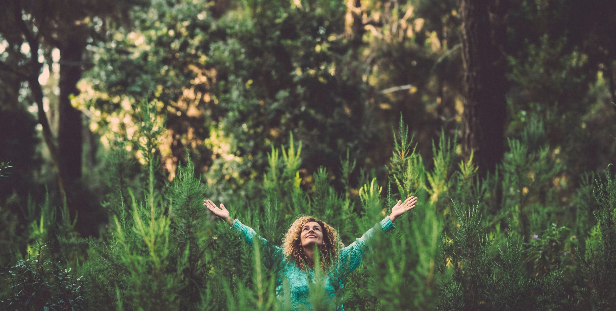 Woman with curly hair stands among tall greenery, arms raised joyfully, surrounded by a lush forest backdrop.