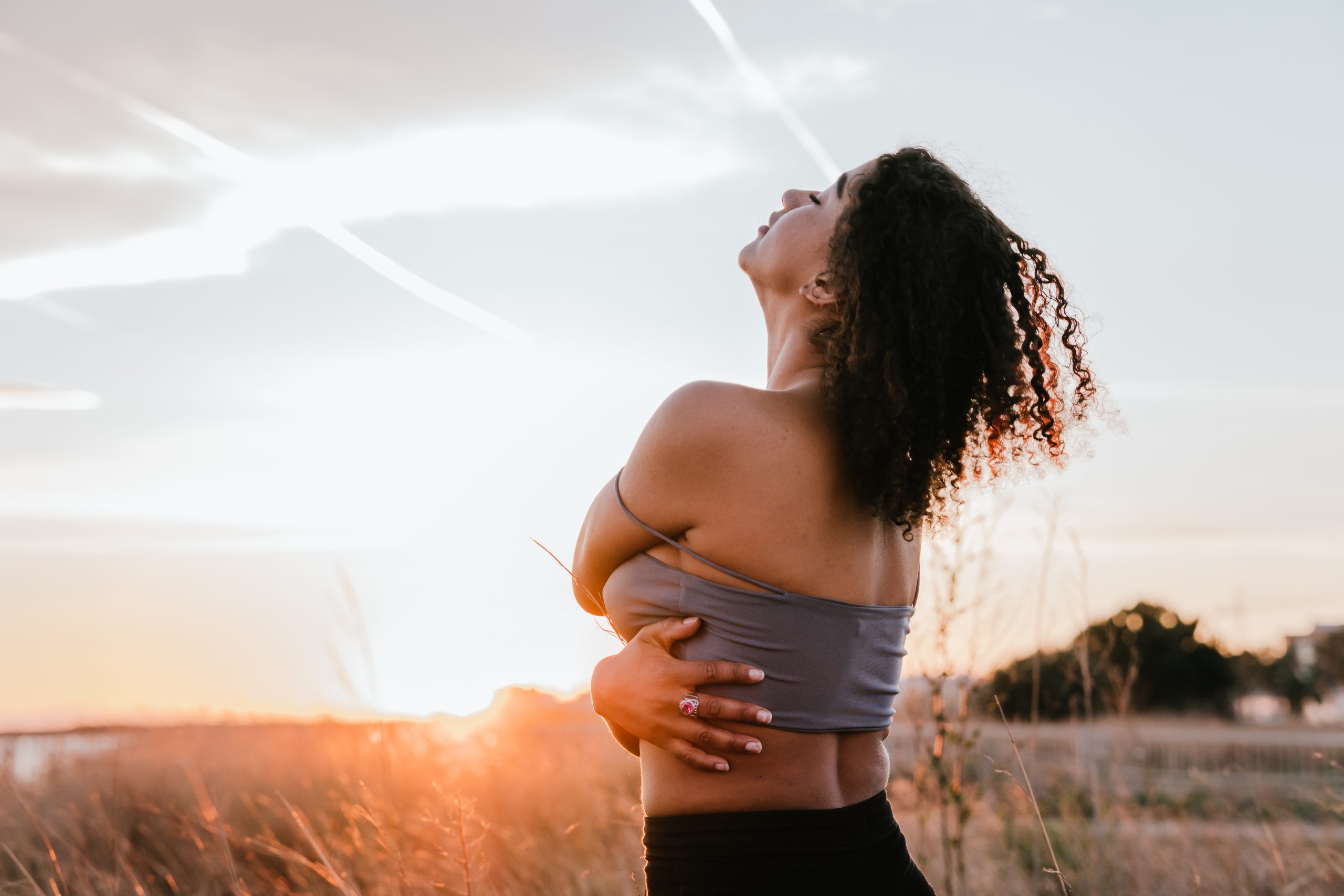 Woman with curly hair stands in a field at sunset, embracing herself with a serene expression against a colorful sky.