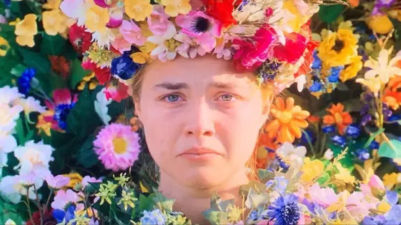 A woman with a floral crown made of vibrant flowers, surrounded by a colorful floral backdrop, looks somber.
