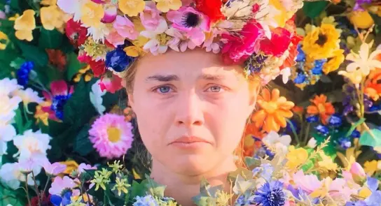 A woman with a floral crown made of vibrant flowers, surrounded by a colorful floral backdrop, looks somber.