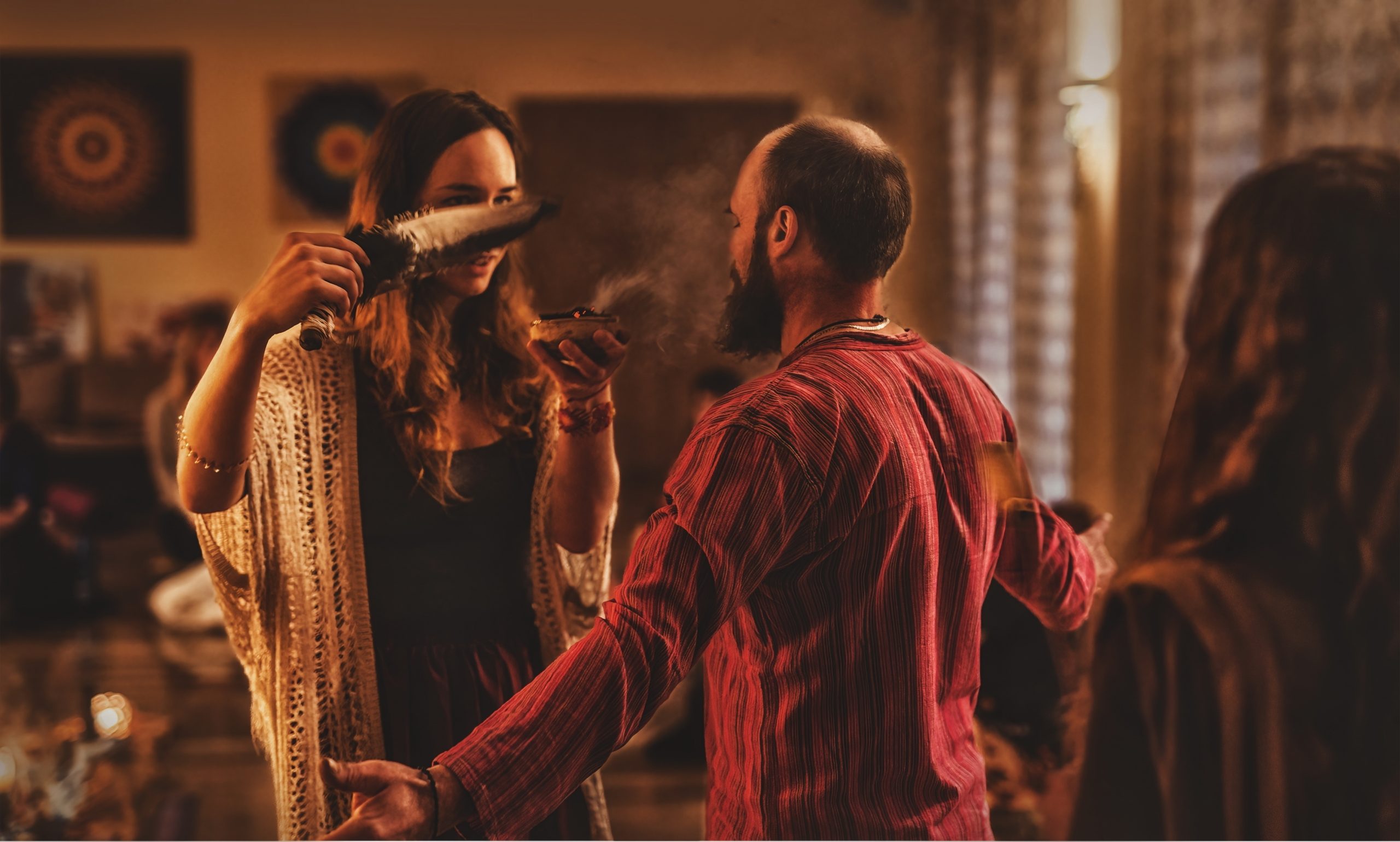 A woman in a flowing cardigan smudges a man with smoke from a bowl, surrounded by a warm, inviting atmosphere.