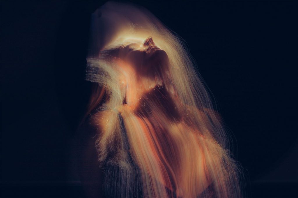 A woman with flowing hair tilts her head back, surrounded by a soft, ethereal glow against a dark background.