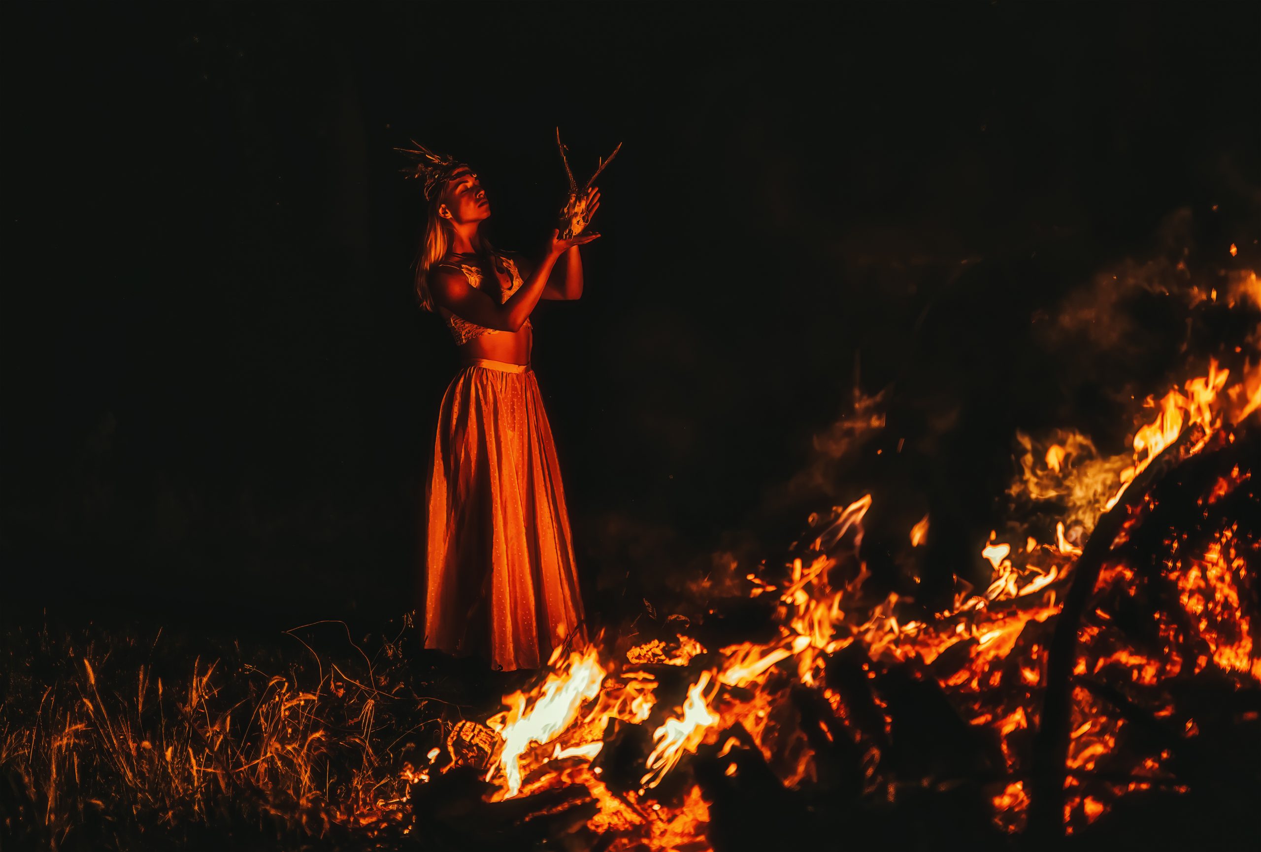 A woman in a flowing skirt stands by a large bonfire, holding twigs, illuminated by the flames against a dark backdrop.
