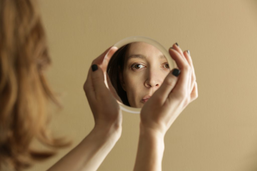 A woman gazes into a round mirror, her reflection showing a contemplative expression against a beige background.