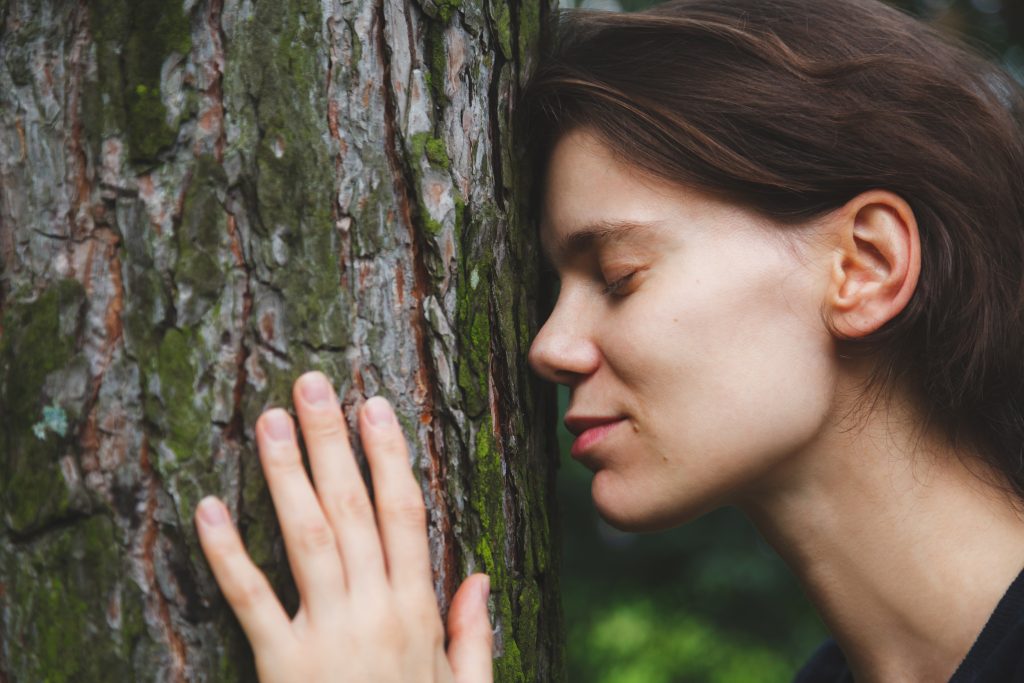 Woman gently resting her cheek against a textured tree trunk, eyes closed, with a serene expression.