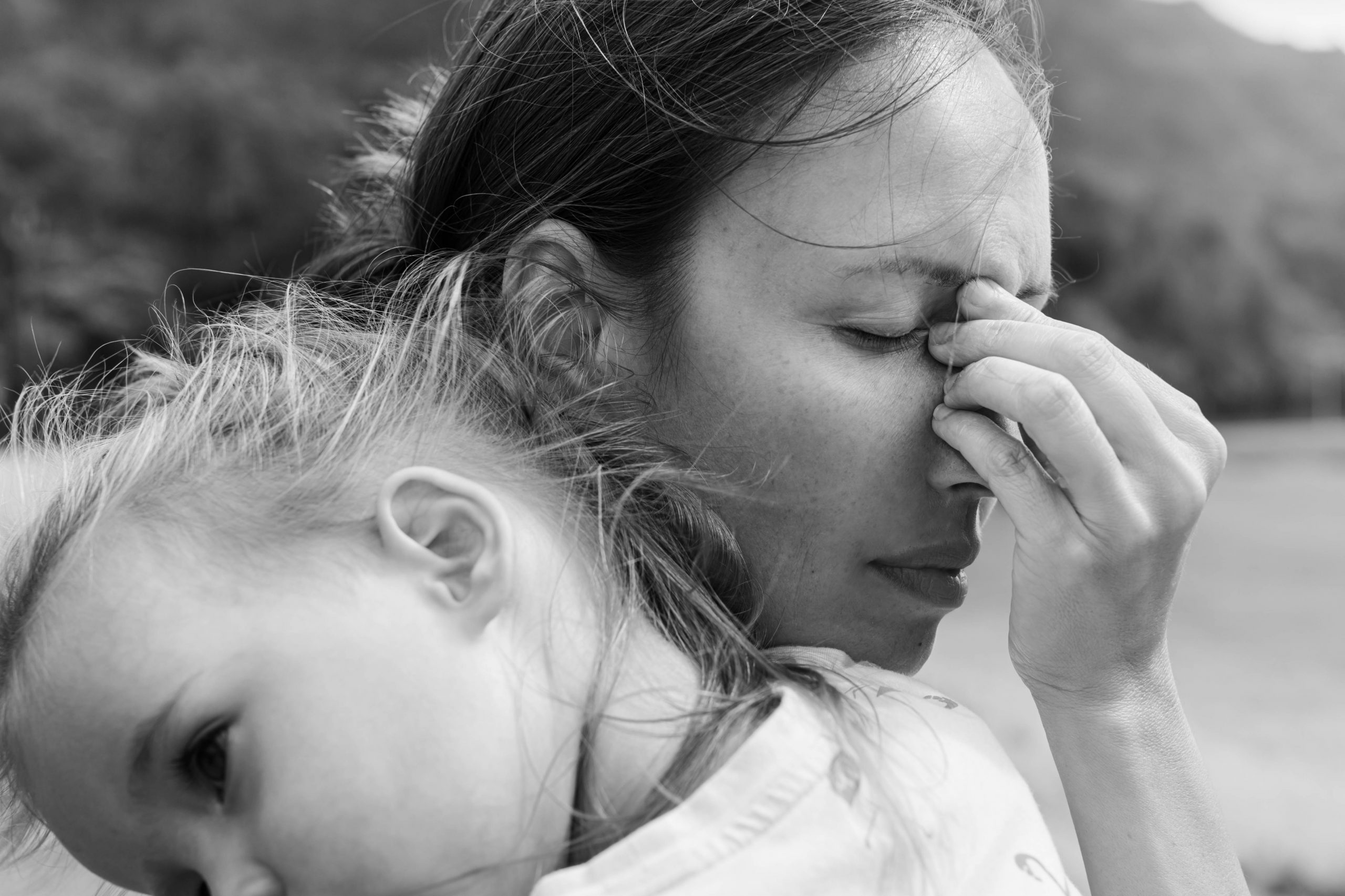 A woman holds a child on her back, visibly stressed, with her hand on her forehead, set against a blurred natural background.
