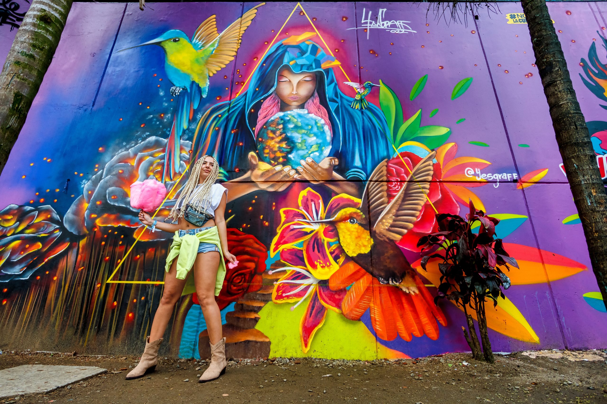 Woman with long braids poses in front of a vibrant mural featuring a cloaked figure holding the Earth, surrounded by color...