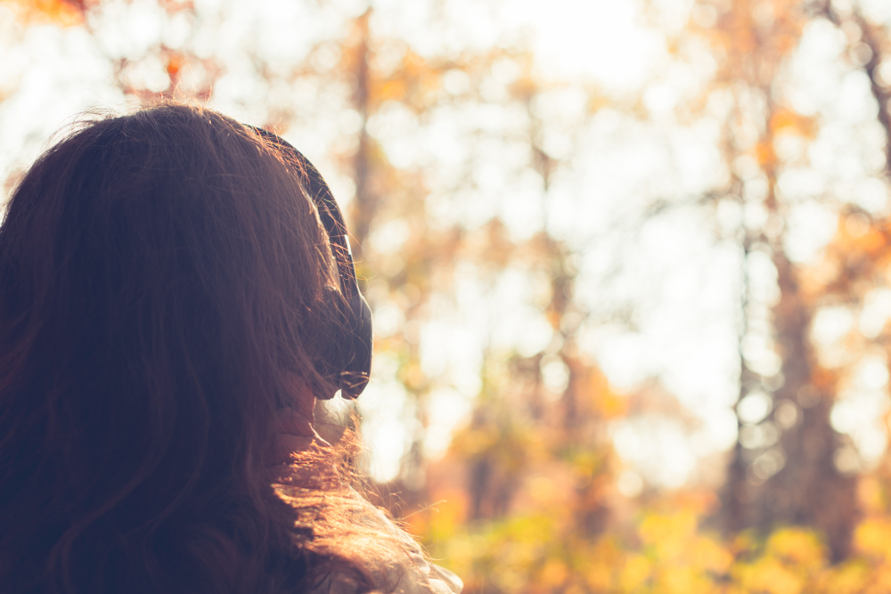 Woman with long hair stands in a sunlit forest, surrounded by autumn foliage, gazing into the distance.