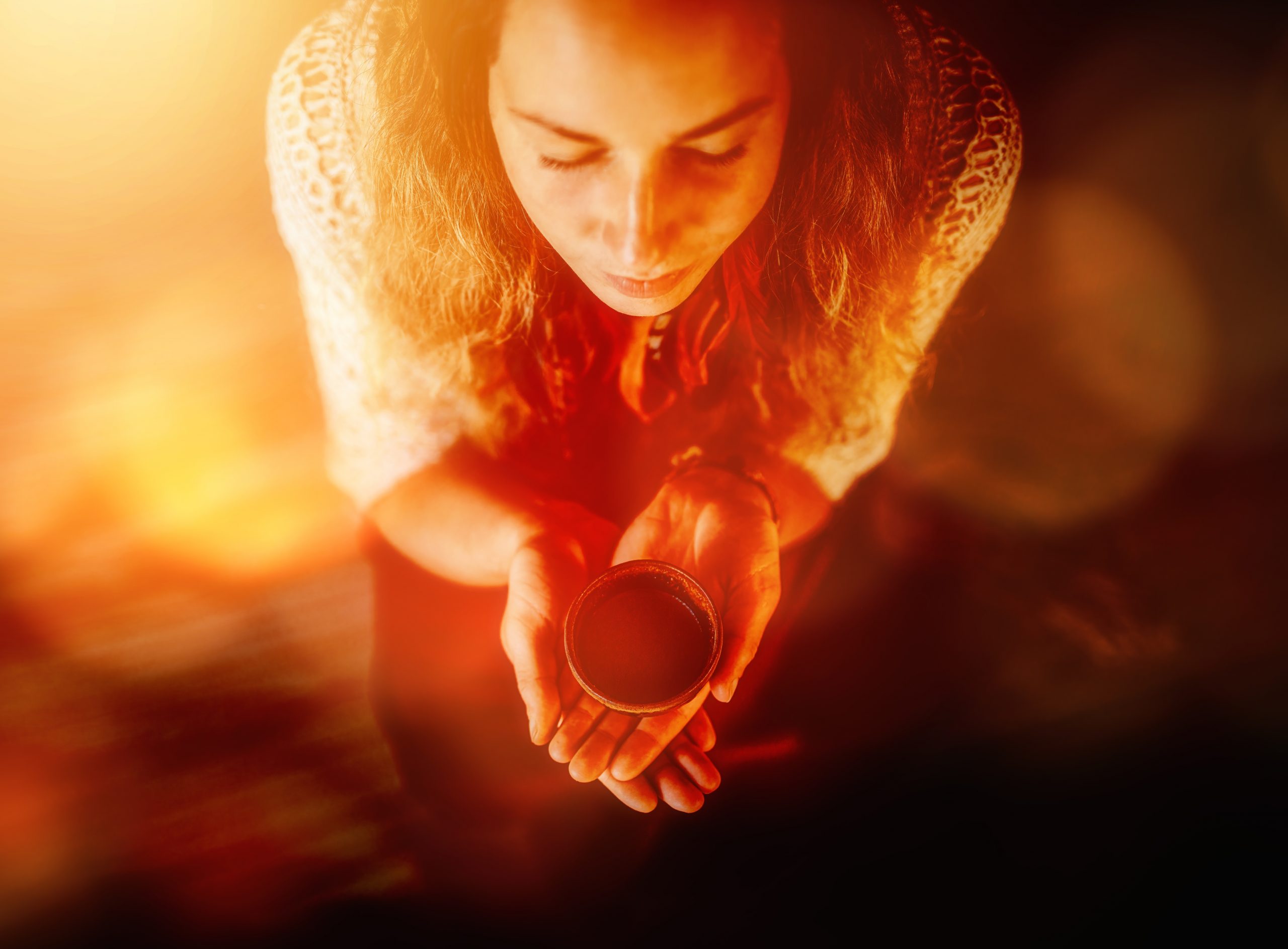 A woman with long, wavy hair holds a small bowl close to her face, surrounded by warm, glowing light.