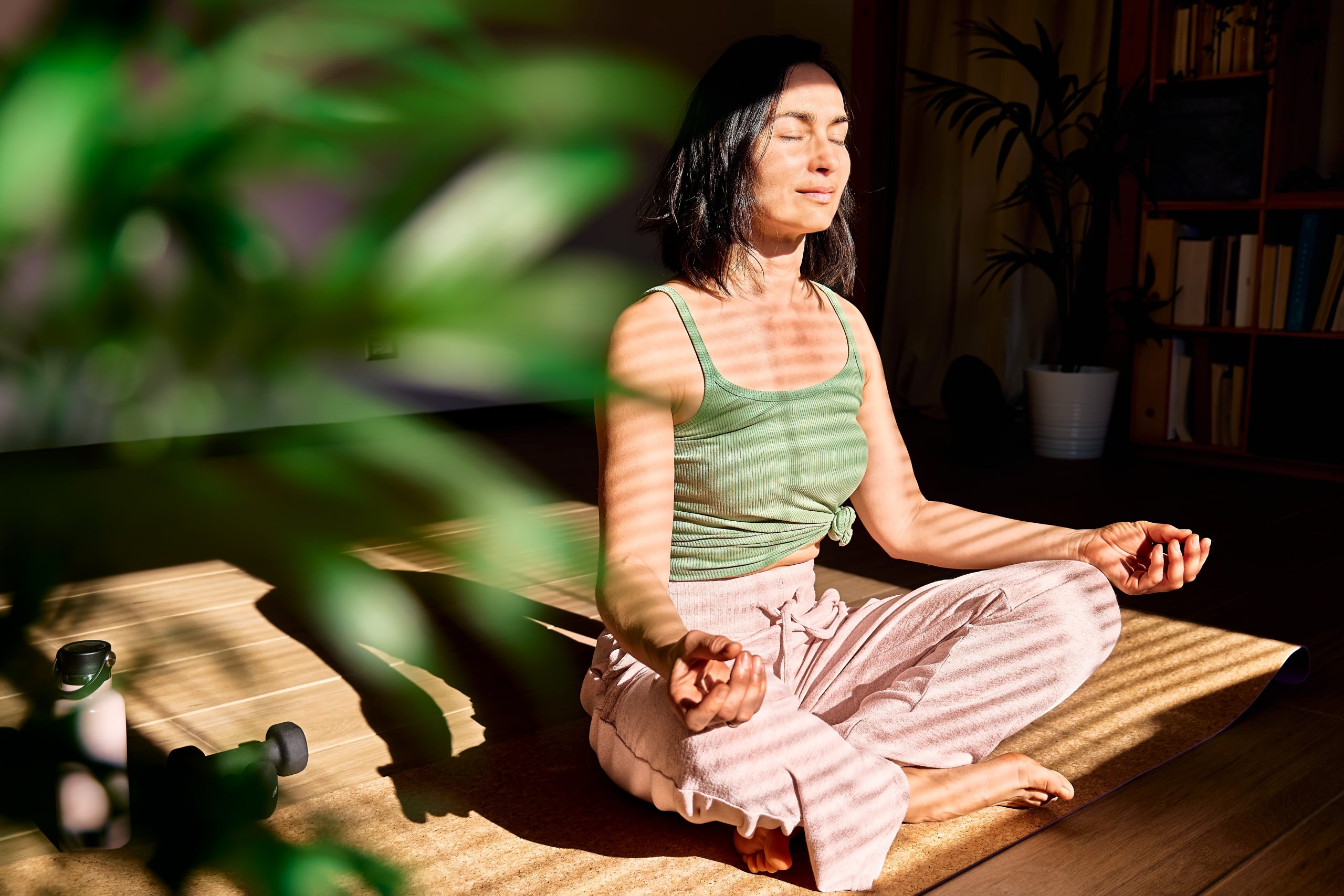 A woman meditates in a sunlit room, surrounded by plants, with shadows creating stripes across her serene face.