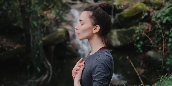 Woman meditating outdoors, eyes closed, hands on her chest, with a serene waterfall and lush greenery in the background.