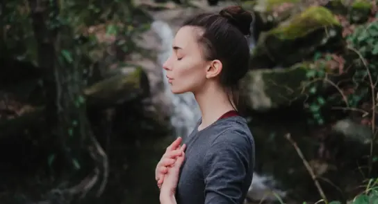 Woman meditating outdoors, eyes closed, hands on her chest, with a serene waterfall and lush greenery in the background.