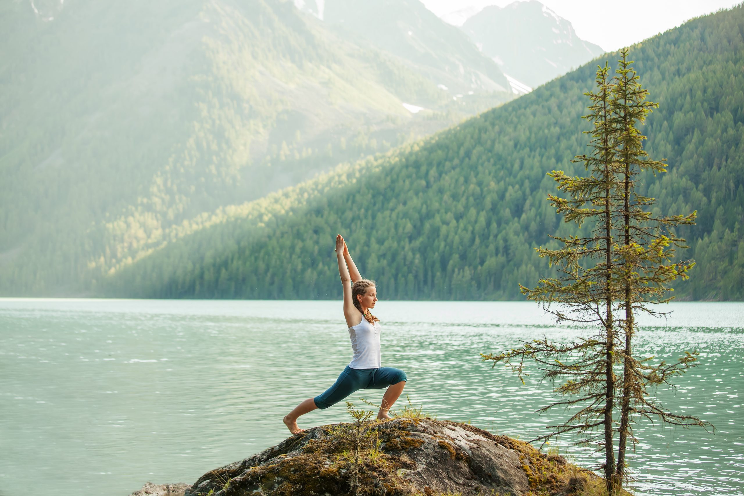 Woman practicing yoga in warrior pose on a rock by a serene lake, surrounded by lush mountains and trees.